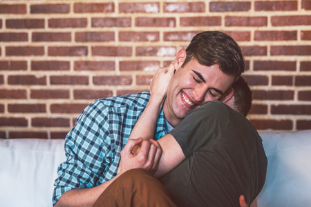 Affectionate young gay couple laughing and hugging each other while sitting together on their sofa at homeの写真素材