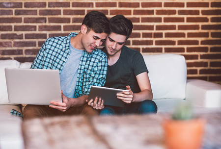 Smiling young gay couple sitting on the sofa at home using a laptop and digital tabletの写真素材