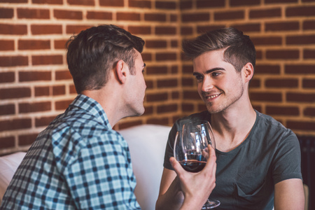 Affectionate young gay couple sitting at home together on their living room sofa talking and drinking red wineの写真素材