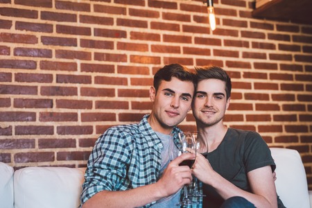 Portrait of an affectionate young gay couple sitting at home together on their living room sofa talking and drinking red wineの写真素材