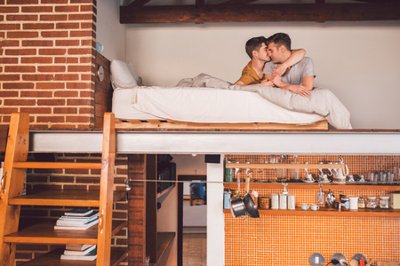 Affectionate young gay couple lying face to face together in their loft bedroom in the morningの写真素材