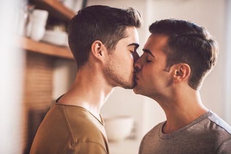 Affectionate young gay couple kissing each other with their eyes closed while standing in their kitchen in the morning