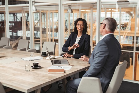 Mature businessman and young work colleague discussing business while sitting together at a table in an office boardroom