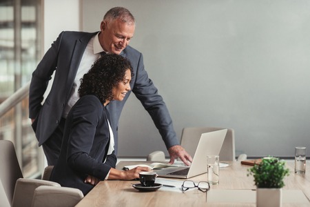 Mature businessman and young work colleague talking together and working on a laptop while working at a table in an office boardroomの写真素材