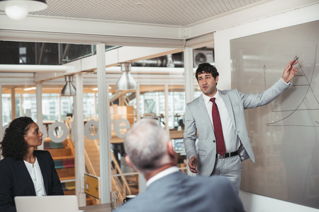 Young businessman standing in a boardroom giving a presentation on a whiteboard to two work colleagues sitting a tableの写真素材