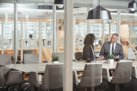 Mature businessman and young work colleague discussing business while sitting together at a table inside a glass office boardroomの写真素材