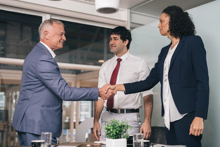 Mature manager shaking hands with a young staff member while standing in an office boardroom with a...