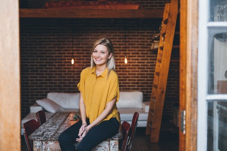 Portrait of an attractive young blonde woman smiling while sitting on a table her modern loft apartmentの写真素材