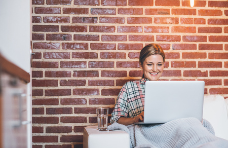 Attractive young blonde woman smiling and using a laptop while sitting on her sofa wrapped in a blanketの写真素材