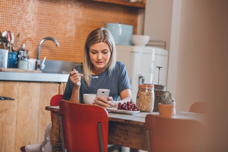 Attractive young blonde woman sitting at a table in her kitchen using a cellphone while eating a...