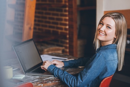 Portrait of an attractive young blonde woman looking over her shoulder while sitting at a table in her loft apartment using a laptopの写真素材