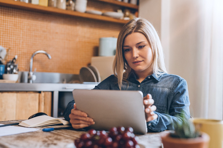 Attractive young blonde woman sitting at a table in her apartment kitchen using a digital tabletの写真素材