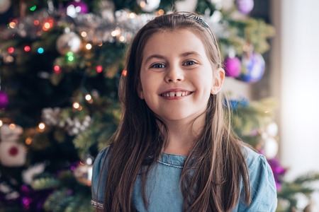 Portrait of an adorable little girl smiling while standing in front of a Christmas tree during the holidaysの写真素材