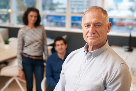 Confident manager standing in an office with staff behind himの写真素材