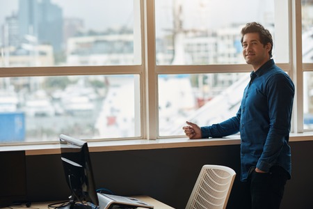 Smiling businessman standing by office windows overlooking the cityの写真素材