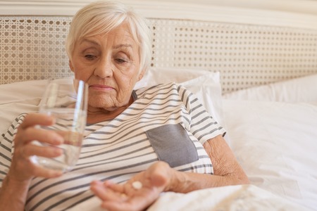 Unhappy senior woman lying in bed taking medicineの写真素材
