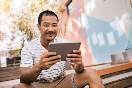 Smiling Asian man sitting on stairs outside using a tabletの写真素材