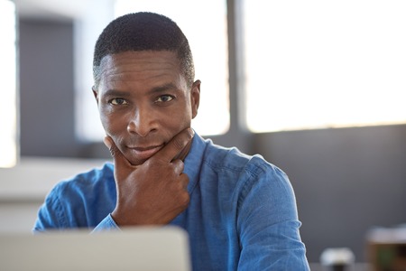 Portrait of a focused young African businessman sitting confidently at his desk in a large modern...