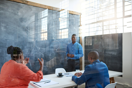 Focused young African businessman taking questions from colleagues while giving a presentation on a chalkboard in a modern officeの写真素材