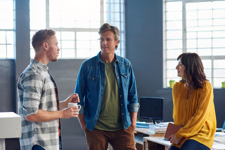 Three casually dressed young office coworkers smiling and talking together while standing in a large modern officeの写真素材