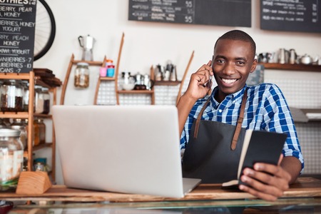 Smiling young African entrepreneur standing at the counter of his cafe talking on a cellphone and reading from a notebook while using a laptopの写真素材