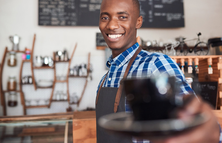 Portrait of a smiling African barista standing at the counter of a trendy cafe offering up a fresh cup of cappuccino の写真素材