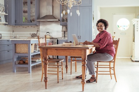 Smiling African woman sitting in her kitchen using a laptopの写真素材