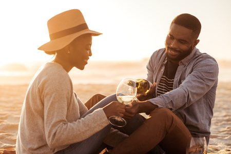 Smiling African couple drinking wine at the beach at sunsetの写真素材
