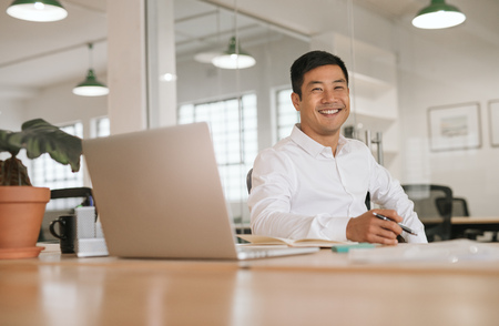 Smiling young Asian businessman working alone at his office deskの写真素材