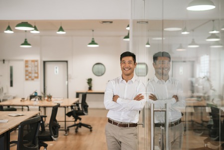 Smiling Asian businessman leaning against a glass wall at workの写真素材
