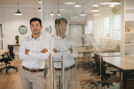 Asian businessman leaning against a glass wall in an officeの写真素材