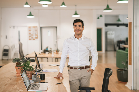 Smiling Asian businessman standing by his desk in an officeの写真素材