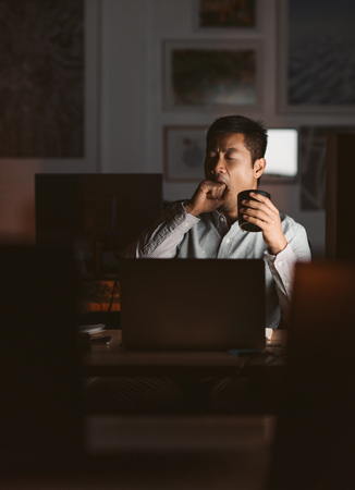 Exhausted businessman yawning and drinking coffee in a dark offi