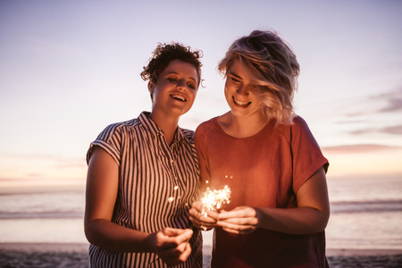 Laughing female friends playing with sparklers during a beach sunsetの写真素材