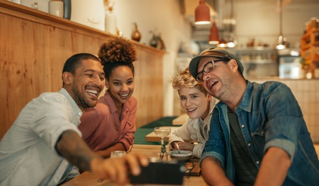 Smiling young friends sitting in a bistro taking selfies together