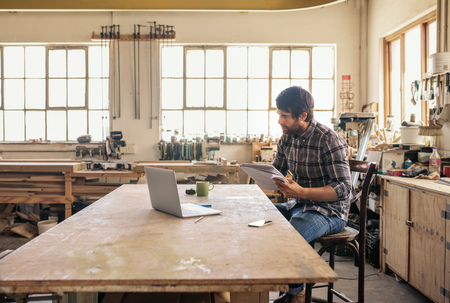 Woodworker using a laptop and reading notes in his workshopの写真素材