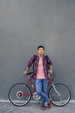 Young man standing with his bike against a gray wallの写真素材