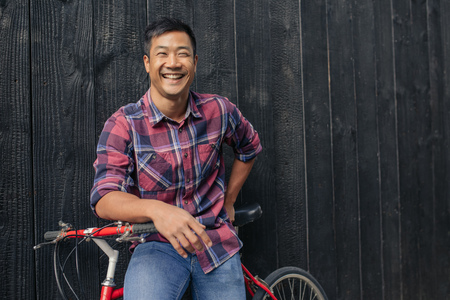 Portrait of a smiling young Asian man in a plaid shirt sitting on his bike leaning against a dark wall in the cityの写真素材