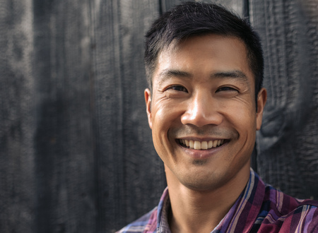 Cheerful young Asian man leaning against black wooden wallの写真素材
