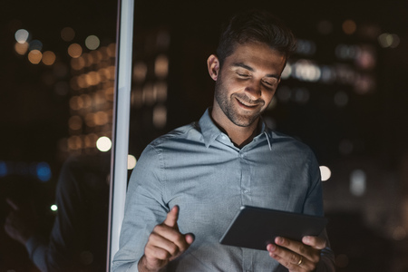 Smiling businessman standing in his dark office using a tabletの写真素材