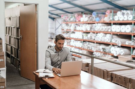 Smiling manager sitting in his warehouse office using a laptopの写真素材