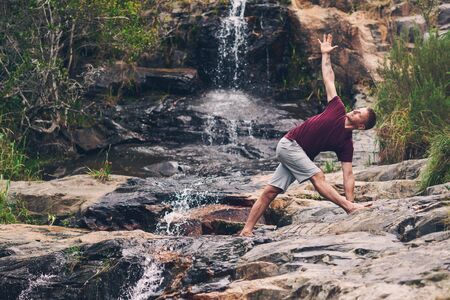 Fit man doing the triangle pose by a waterfallの写真素材