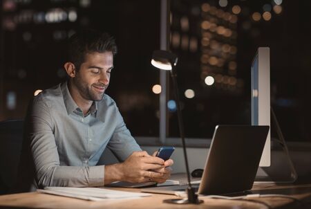 Smiling young businessman reading a text message while working lateの写真素材