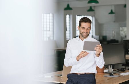 Smiling businessman leaning on an office desk using a tabletの写真素材