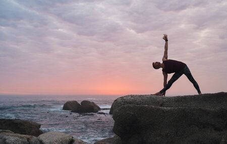 Man doing the triangle pose by the ocean at duskの写真素材