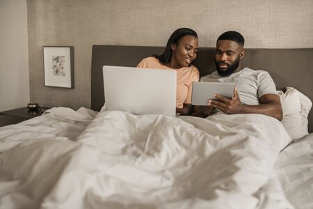 African American couple lying in bed checking their social mediaの写真素材