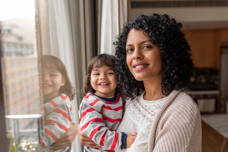 Smiling mother and daughter standing by their living room windowの写真素材