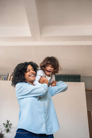 Mother playfully holding her laughing little daughter over her shoulder while having fun together at homeの写真素材