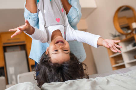 Laughing little daughter being held upside down by her mom over their sofa while playing together at homeの写真素材