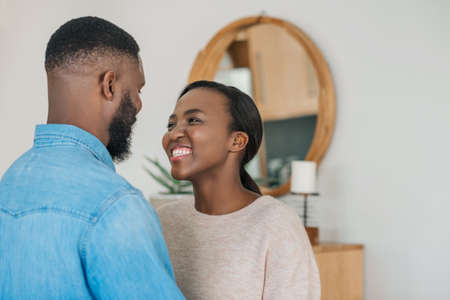 Young African American woman smiling and looking at her husband while standing arm in arm together in their modern apartmentの写真素材